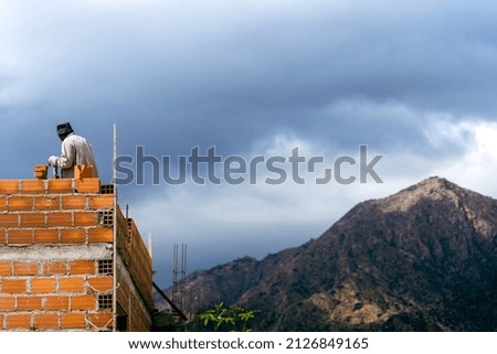 A man building a brick wall with a mountain and a cloudy sky on the background