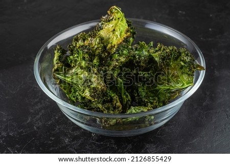 Healthy chips baked in the oven from kale in a transparent bowl. Dark background.