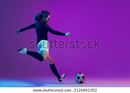 Dynamic portrait of female soccer player practicing with football ball isolated on purple studio background in neon light. Concept of sport, action, motion, fitness. Young spotive girl training