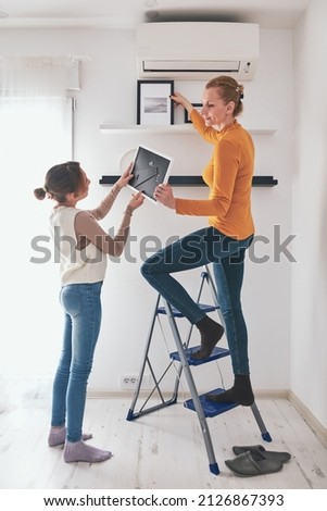 Mother and daughter on the ladder hanging pictures and photos on the shelf and wall at home.