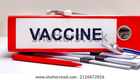 On the desktop is a stethoscope, documents, a pen, and a red file folder with the text VACCINE. Medical concept
