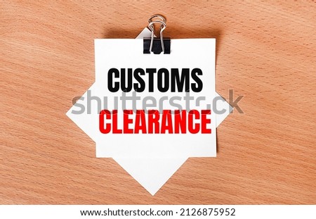 On a wooden table under a black paper clip lies a sheet of white paper with the text CUSTOMS CLEARANCE