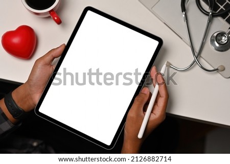 Top view of hands holding a blank white screen tablet and a pen leaning on the white table with a heart model, laptop and a stethoscope on the sides for medical, health care and technology concept.