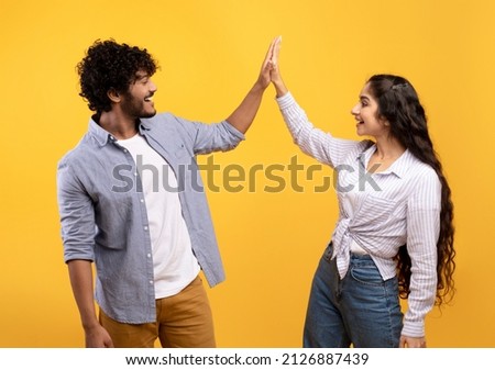 Common success. Young excited indian couple giving high-five, clapping hands, greeting each other and smiling, standing over yellow studio background