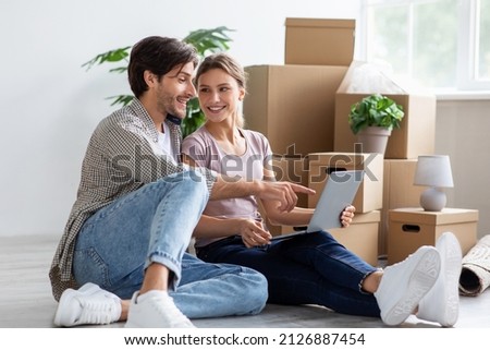 Happy millennial caucasian female and guy in casual looking for ideas for new interior at computer sitting among cardboard boxes in room. Moving, buying apartment, rent home and relocation together