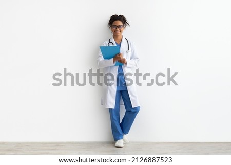 Friendly female black doctor in glasses holding clipboard looking at camera standing leaning on wall, lady wearing blue uniform, coat and stethoscope, full body length, white studio background Royalty-Free Stock Photo #2126887523