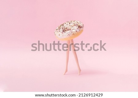 Small ring donut with white glaze and colorful crumbs with plastic model woman toy legs walking on a pink pastel background. Minimal design and concept.