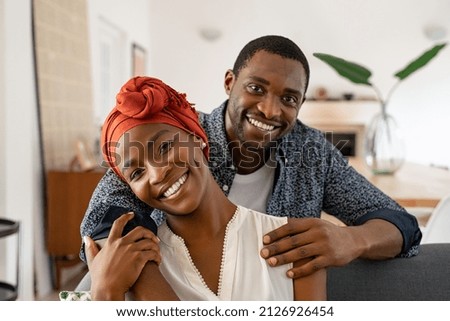 Portrait of mid adult happy african american couple hugging at home while sitting on couch. Portrait of mature man with his woman wearing traditional turban looking at camera. Black mid adult couple.  Royalty-Free Stock Photo #2126926454