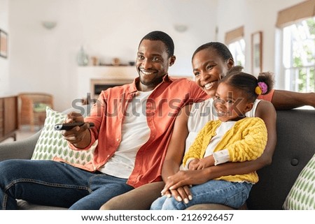 Relaxed african american family watching TV together. Happy mature father changing television channel using remote control with daughter sitting on mother's lap at home. Black family watching movie . Royalty-Free Stock Photo #2126926457