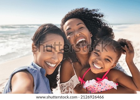 Portrait of smiling young african american woman with child taking selfie at beach with her best friend. Cheerful multiethnic gay couple enjoying at beach with daughter during summer holiday.  Royalty-Free Stock Photo #2126926466