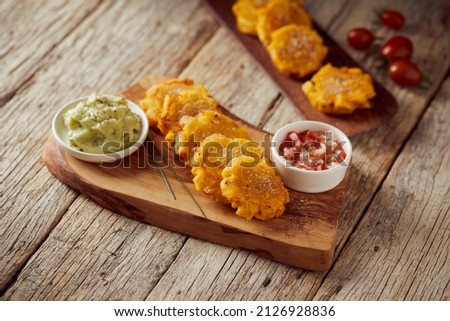 Patacones or tostones, typical Ecuadorian appetizer that consists on fried green plantain slices. It’s accompanied with guacamole and served on a traditional plate with a wooden and rustic background. Royalty-Free Stock Photo #2126928836