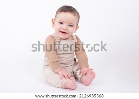 Adorable baby boy wearing beige overalls sitting on white background looking at camera and smiling.  Royalty-Free Stock Photo #2126935568