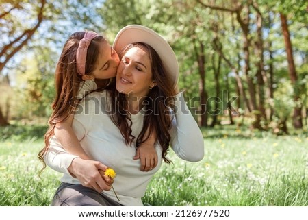 Girl kisses mother on cheek hugging while sitting on grass in spring park giving flower. Family relaxing outdoors. Mothers day. Space