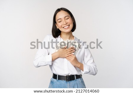 Happy asian woman hugging money dollars and smiling satisfied, standing over white background