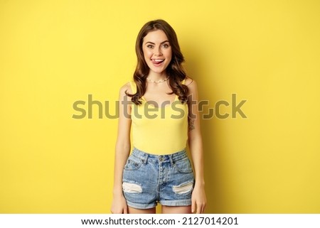 Stylish young brunette woman showing her white healthy teeth, smiling and showing tongue, winking enthusiastic, standing over yellow background