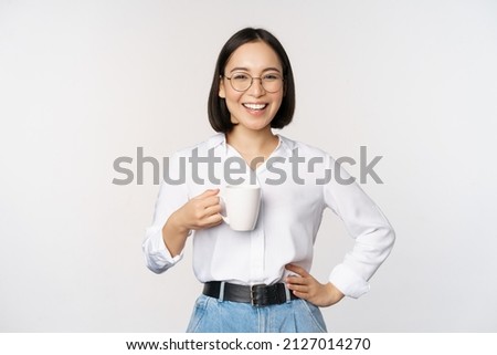 Happy young energetic asian woman smiling, drinking, holding cup mug of coffee, standing confident against white background Royalty-Free Stock Photo #2127014270