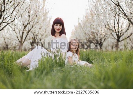 Happy pretty daughter sitting with mother among beautiful spring nature, smiling and looking at camera. Caucasian family of two resting during free time at blooming garden.