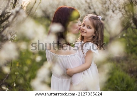 Caucasian caring mother embracing and kissing her lovely daughter that holding on hands among spring garden with blooming apple trees. Concept of family, relations and love.