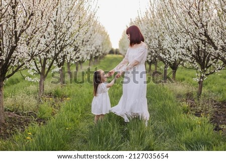 Happy lovely mother holding hands with her little cute daughter while playing together among spring garden. Caucasian family of two in white dresses enjoying leisure time on fresh air.