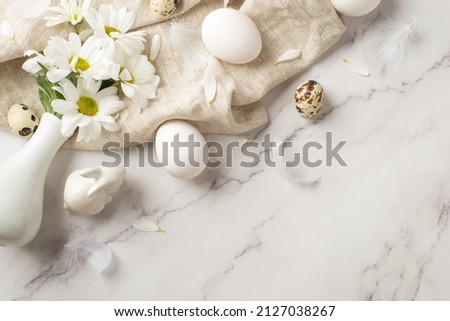 Top view photo of the white vase with daisies scattered petals and feathers few eggs and ceramic rabbit on the textured cloth on isolated marble empty background
