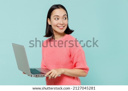 Young smiling happy woman of Asian ethnicity 20s in pink sweater hold use work on laptop pc computer look aside on workspace area isolated on pastel plain light blue color background studio portrait. Royalty-Free Stock Photo #2127045281