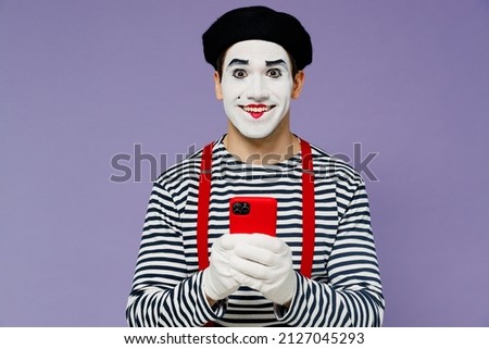 Smiling fun amazing marvelous ecstatic young mime man with white face mask wears striped shirt beret hold in hand use mobile cell phone isolated on plain pastel light violet background studio portrait Royalty-Free Stock Photo #2127045293