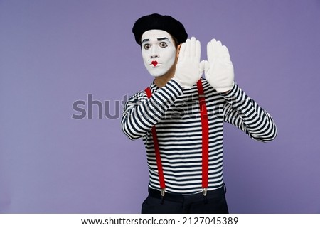 Fun blithesome charismatic vivid young mime man with white face mask wears striped shirt beret gloves looking from behind raised hands isolated on plain pastel light violet background studio portrait Royalty-Free Stock Photo #2127045389
