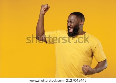 Young exultant happy black man 20s in bright casual t-shirt doing winner gesture celebrate clenching fists say yes isolated on plain yellow color background studio portrait. People lifestyle concept