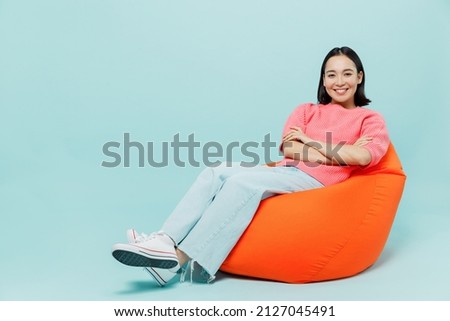 Full body young smiling cheerful happy woman of Asian ethnicity 20s wear pink sweater sit in bag chair look camera isolated on pastel plain light blue color background studio People lifestyle concept