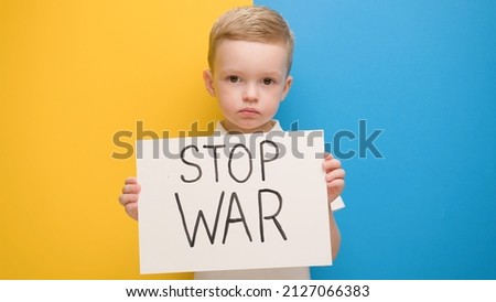 Portrait serious blond 4-year-old boy protesting war raises banner with inscription Stop War at blue yellow flag of country Ukraine. Call to stop war, child against war, crisis in Ukraine. Royalty-Free Stock Photo #2127066383