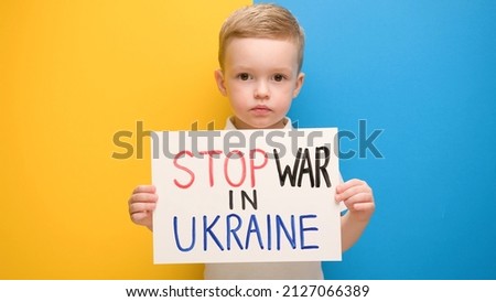 Portrait Blond little boy calls to Stop war in Ukraine, raises banner with inscription stop the war in Ukraine standing on blue-yellow studio background. No war, stop war, russian aggression. Royalty-Free Stock Photo #2127066389