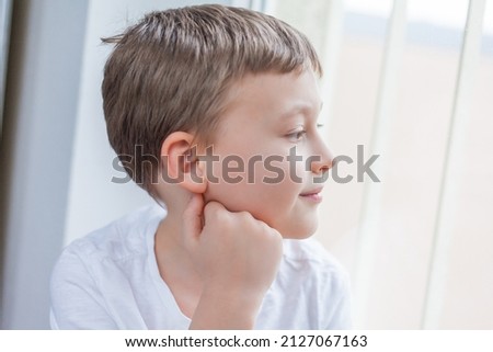 Little boy 7 years old in a white T-shirt in a bright room looks out the window. Emotions. Home.