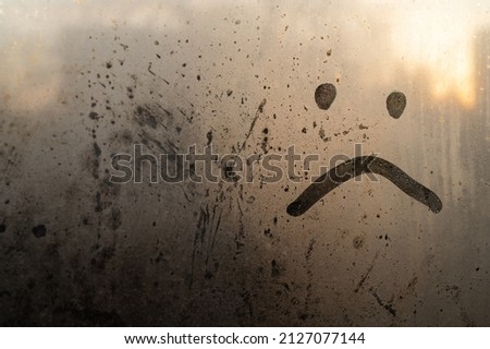 upset smiley, sad, painted on the window over a blurred background, dew and drops of water, rain, morning in the warm rays, unhappy when his rainy weather is a beautiful art. Place for inscription.