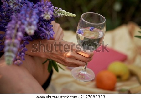 Woman holds a wine glass with purple lupinus at sunset. Summer picnic. Wellness, natural concept. Adaptogenic ayuverdic drink concept. Conscious consumption. Extra wide banner. Purple place for text