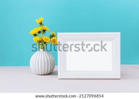 White picture frame mockup with wild yellow dandelion flowers in vase. Empty frame mock up for presentation design. White template on cian background