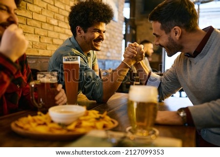 Happy Lebanese man and his male friend arm wrestling and having fun while drinking beer in a pub. 
