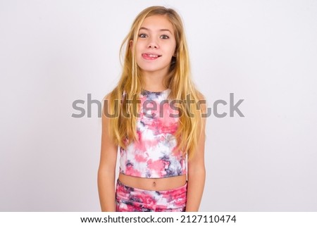 Funny little caucasian kid girl wearing sport clothing over white background makes grimace and crosses eyes plays fool has fun alone sticks out tongue.