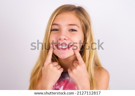 Happy little caucasian kid girl wearing sport clothing over white background with toothy smile, keeps index fingers near mouth, fingers pointing and forcing cheerful smile