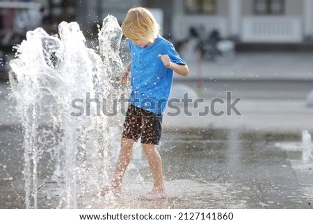 Little boy plays in the square between the water jets in the dry fountain at sunny summer day. Active summer leisure for kids in the city. Royalty-Free Stock Photo #2127141860