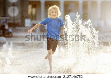 Little boy plays in the square between the water jets in the dry fountain at sunny summer day. Active summer leisure for kids in the city. Royalty-Free Stock Photo #2127146753