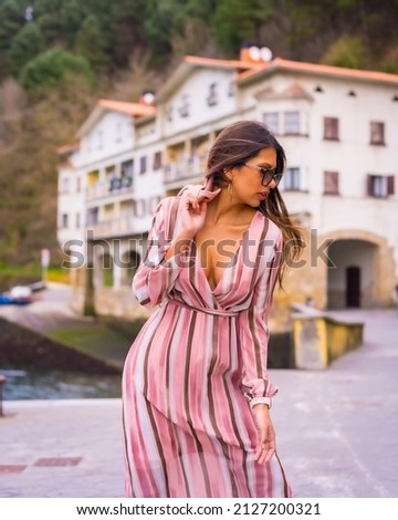 Portrait of a young Latin woman in a pink dress and sunglasses enjoying summer vacations in a beautiful coastal town, posing fashionably by the sea, vertical photo