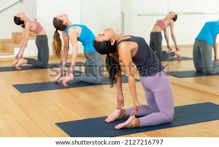 Concentrated hispanic woman performing kneeling back-bending asana Ustrasana (Camel Pose) during group yoga course in fitness studio Royalty-Free Stock Photo #2127216797