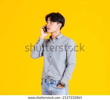 Studio shot of millennial Asian thoughtful doubtful curious male fashion model in stylish fashionable casual outfit standing holding  smartphone on call with friend on yellow background.