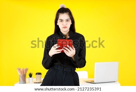 Portrait studio shot of Asian happy beautiful female employee secretary with teeth braces in black outfit sitting smiling on work desk look at camera holding touchscreen tablet on yellow background.