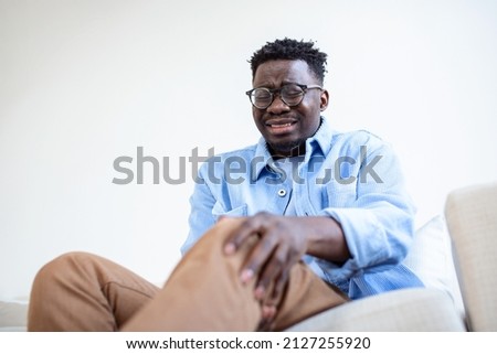 Photo of African man sitting on a sofa in the living room at home and touching his knee by the pain during the day. man massaging his painful knee.