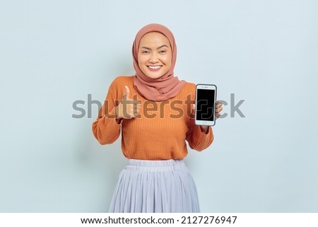Cheerful beautiful Asian woman in brown sweater and hijab, showing blank mobile phone screen, thumb up gesture isolated on white background. Muslim lifestyle concept