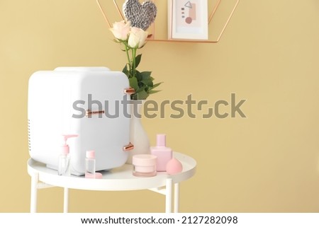 Small refrigerator and cosmetic products on table near color wall