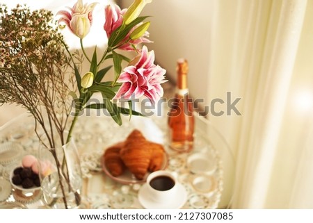 Bouquet of pink lilies. Cup of coffee, croissants and a bottle of wine on a blurred background. Festive breakfast. Light positive photo