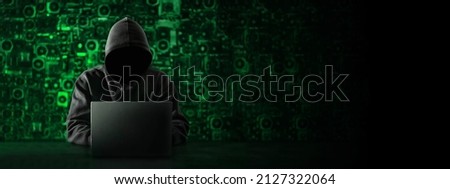 Hacker with a hidden face and with a laptop sits at the table, blue background. There is an hourglass on the hacker's desk. Place for text. High quality photo Royalty-Free Stock Photo #2127322064