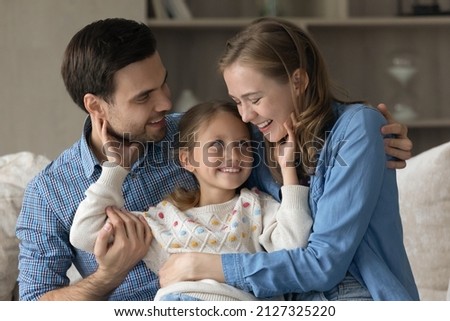 Cheerful family couple of millennial parents cuddling, hugging cute daughter kid on couch in home living room, laughing, having fun, enjoying leisure together. Happy parenthood concept